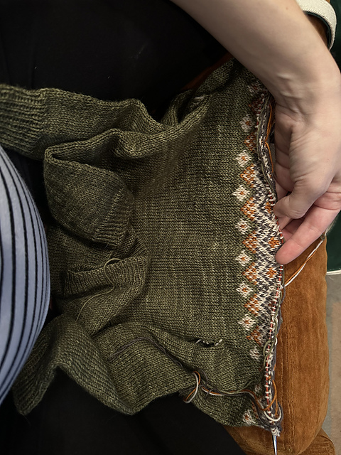 A photo of a green sweater sitting on a woman's lap, with the beginning of cream and orange diamonds at the top. Knitting needles are strung through the top row of stitches. 